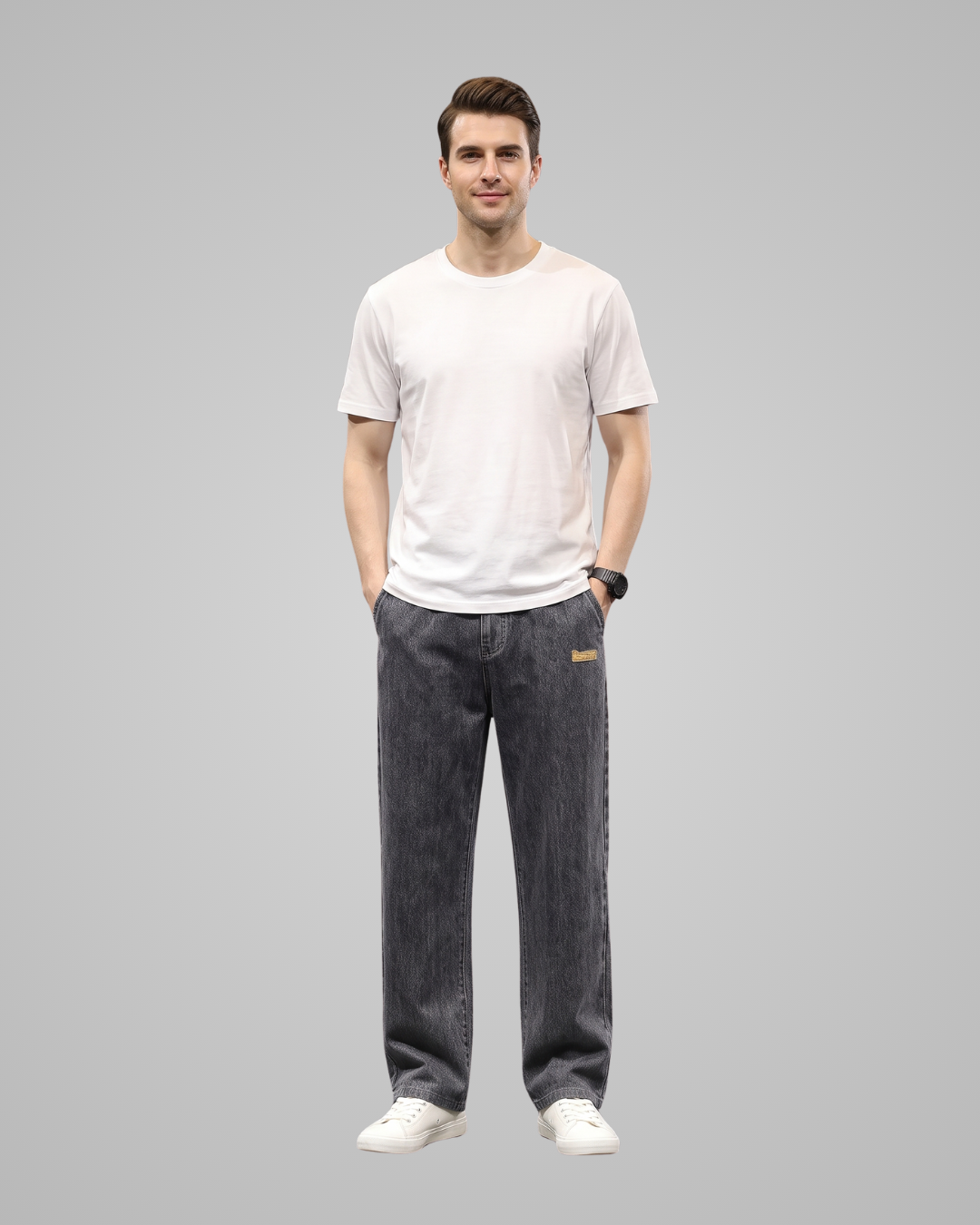 A man wearing a white crew-neck t-shirt, dark gray relaxed-fit jeans with a small gold brand tab on the pocket, and white sneakers, standing against a solid gray background.