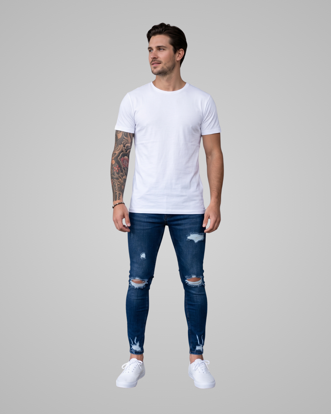 A man modeling a white crew-neck t-shirt, distressed dark blue skinny jeans, and white sneakers against a solid gray background.