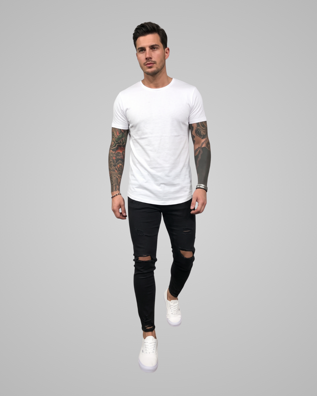 A man modeling a white crew-neck t-shirt, black distressed skinny jeans, and white sneakers against a solid gray studio background.