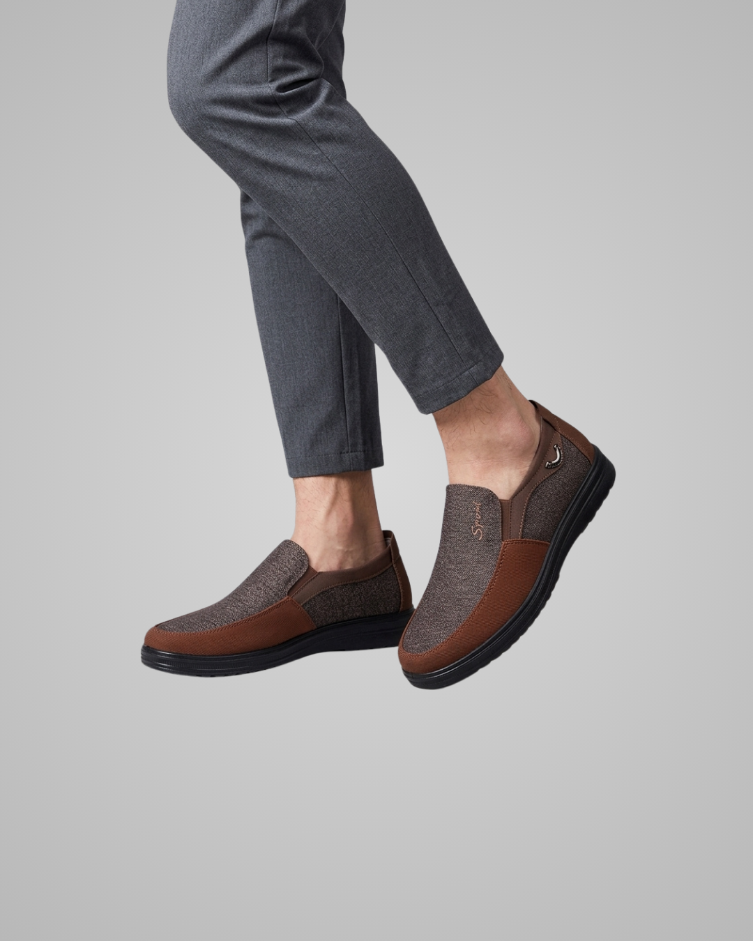 A person wearing brown and gray slip-on loafers with dark gray trousers, shown from the calves down against a plain light gray studio background.