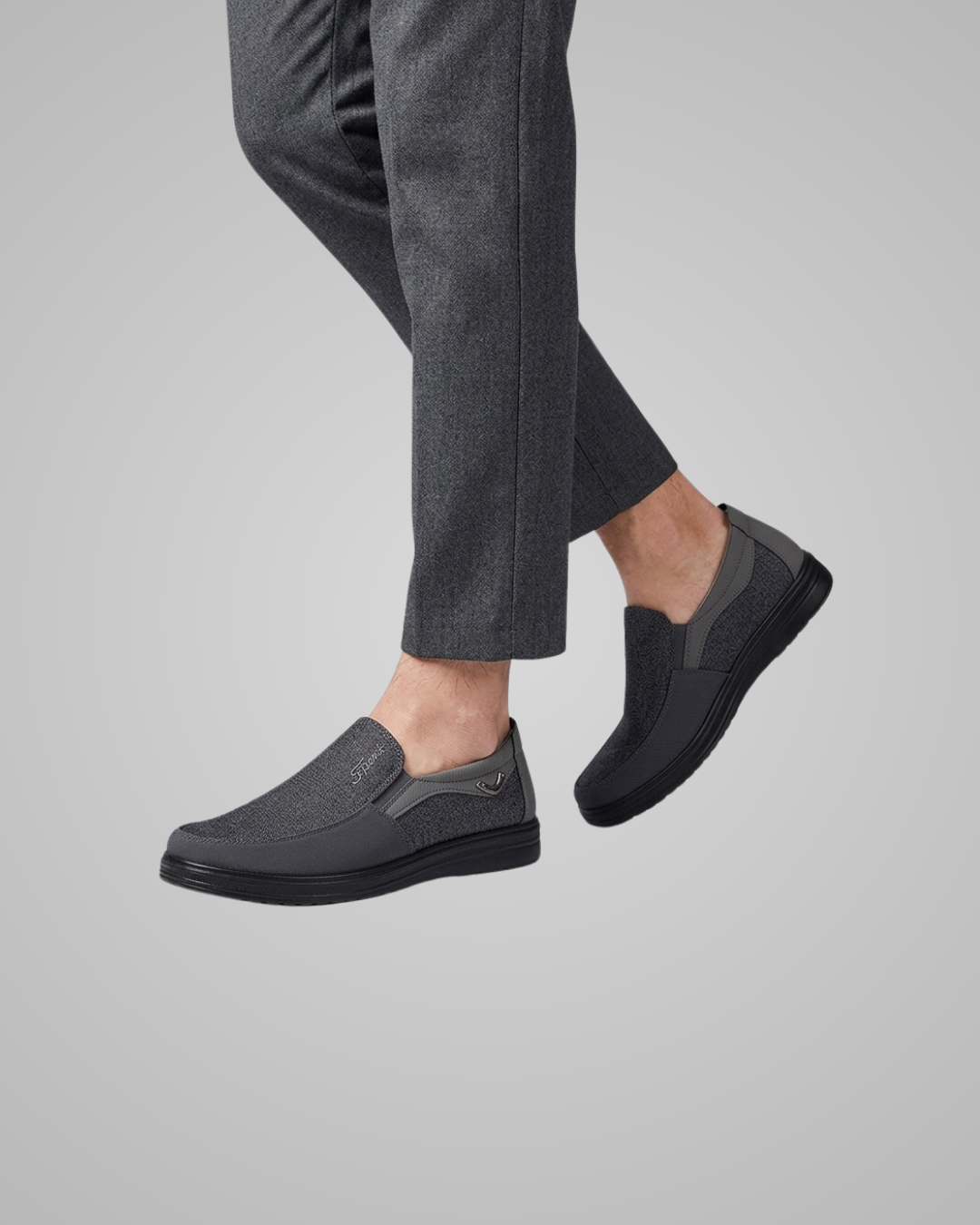 A pair of dark gray slip-on loafers worn with dark gray trousers, shown from the side against a solid light gray background.