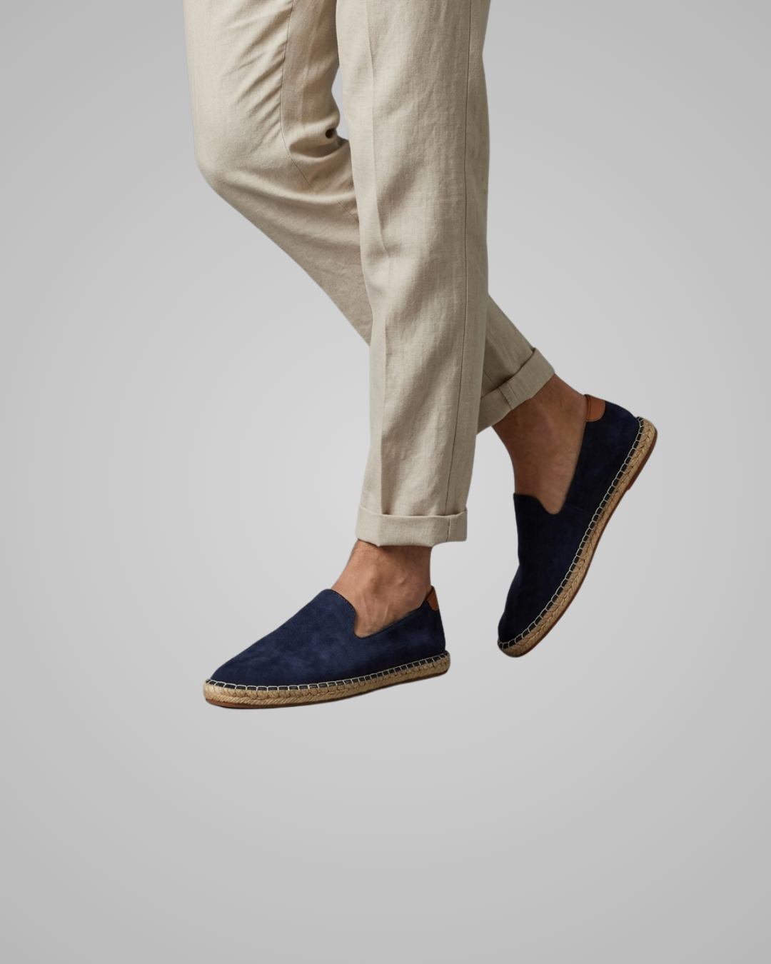 A person wearing navy blue slip-on espadrille-style shoes with tan jute trim and light beige trousers, shown against a plain light gray studio background.