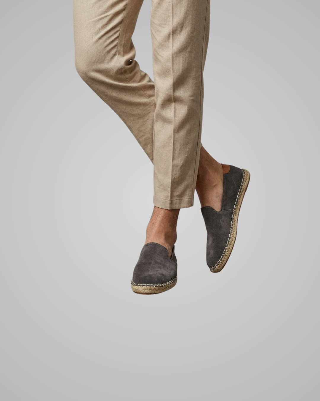 A person wearing beige trousers and dark gray suede espadrille slip-on shoes, shown from the knees down against a light gray studio background.