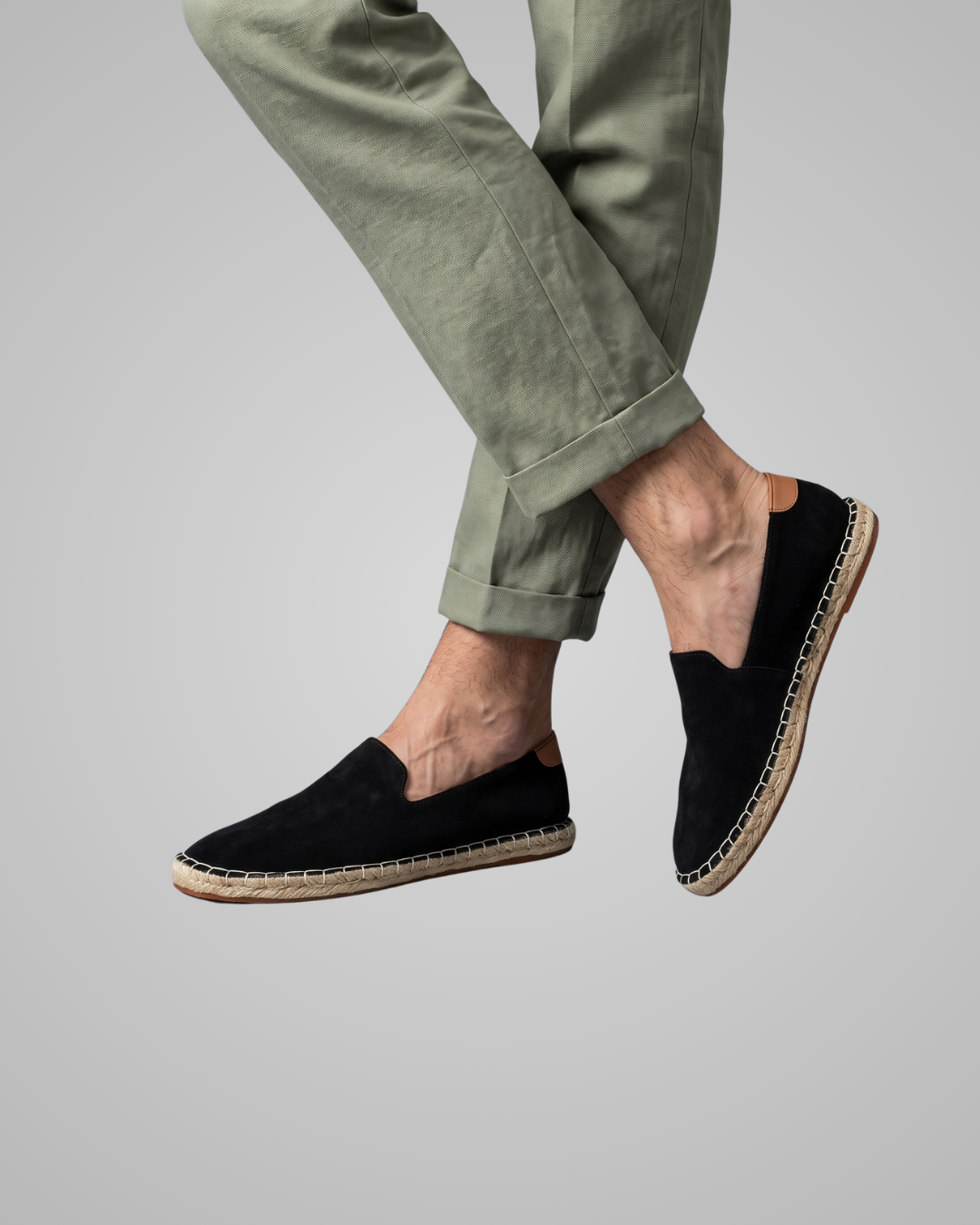 A person wearing black slip-on espadrille-style loafers with natural jute-wrapped soles, shown against a plain light gray studio background. The photo is cropped from the lower legs down, with the pants in an olive green tone.