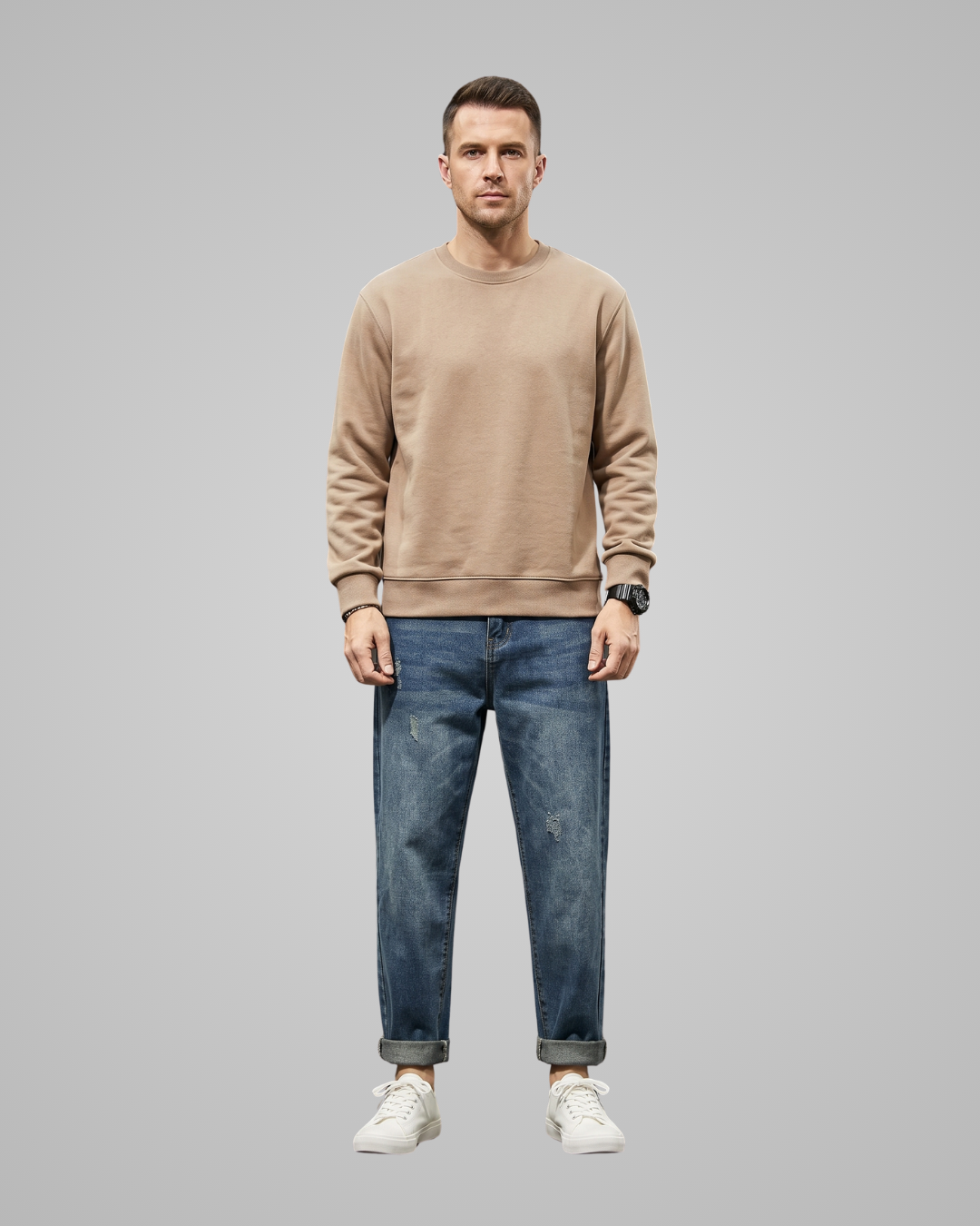 A man stands against a plain light gray studio background wearing a beige crewneck sweatshirt, blue jeans, and white sneakers.