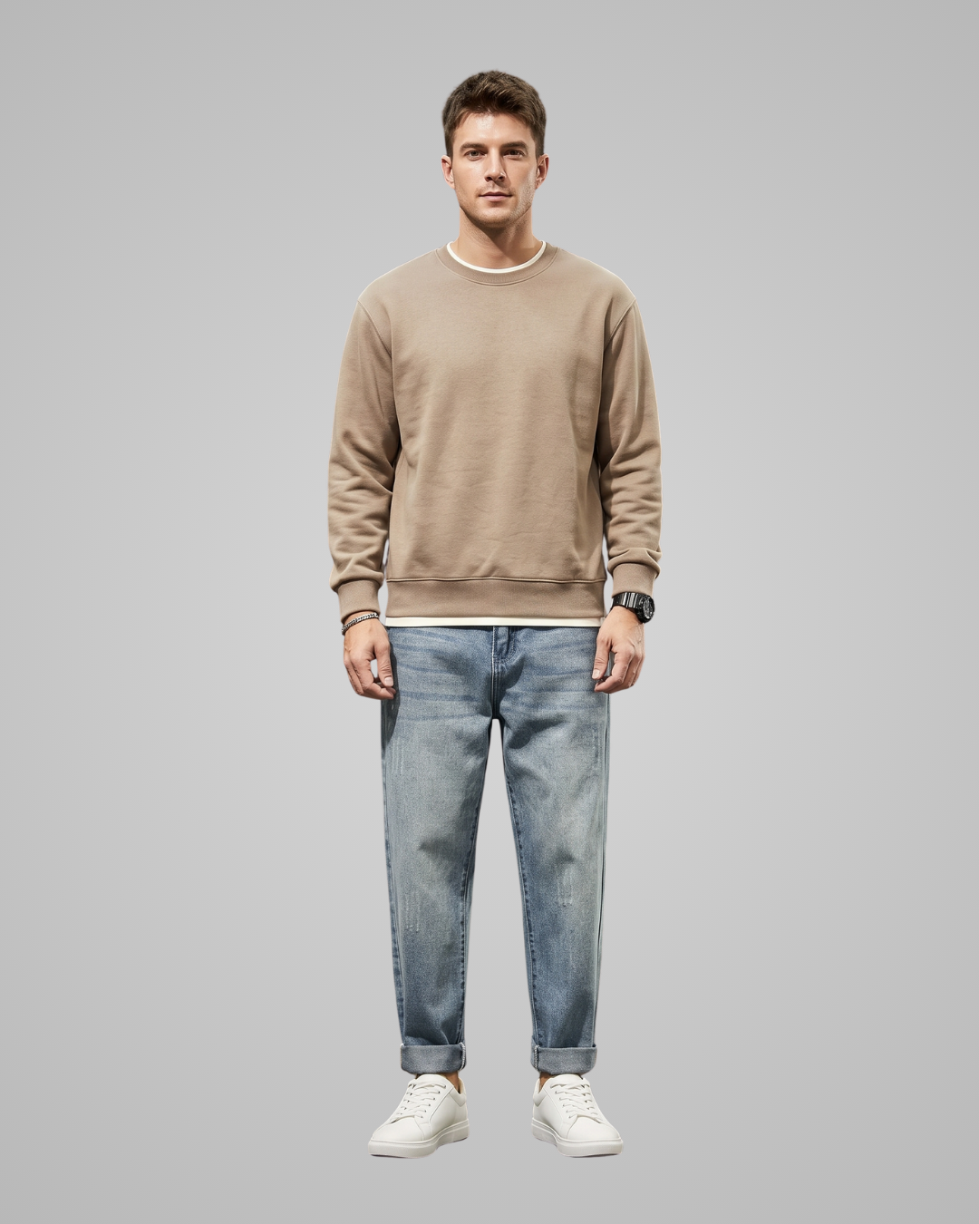 A man modeling a beige crewneck sweatshirt with white trim, paired with light blue jeans and white sneakers, standing against a solid gray background.
