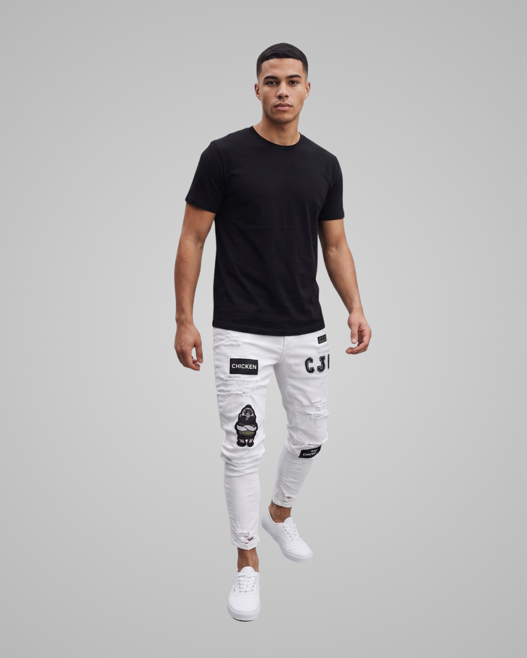 A man modeling a black crew-neck t-shirt and white distressed jeans with black patches and graphics, paired with white sneakers, against a solid gray background.