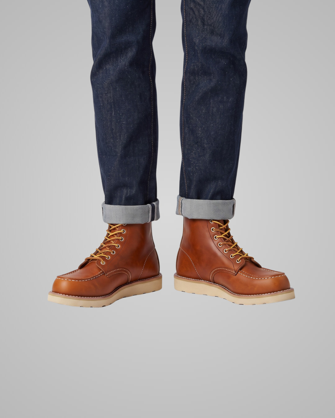 A pair of men's brown leather lace-up boots with light tan soles, shown on a model wearing dark blue cuffed jeans against a solid gray background.