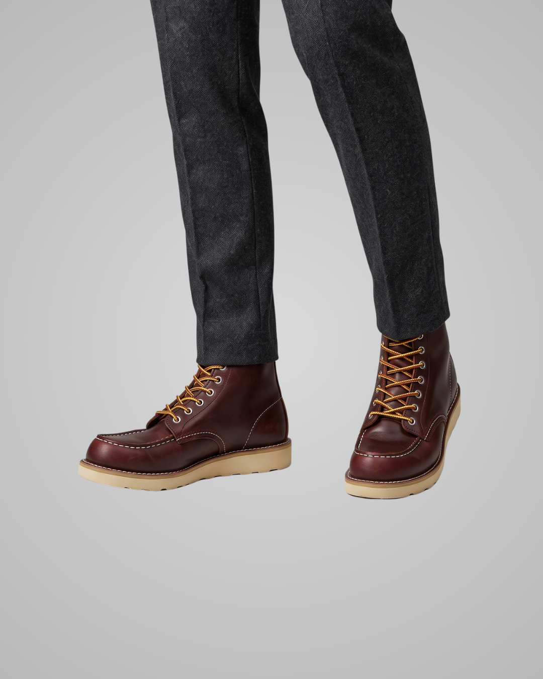 A pair of men's dark brown leather lace-up boots with tan soles and laces, worn with dark gray trousers against a solid gray background.