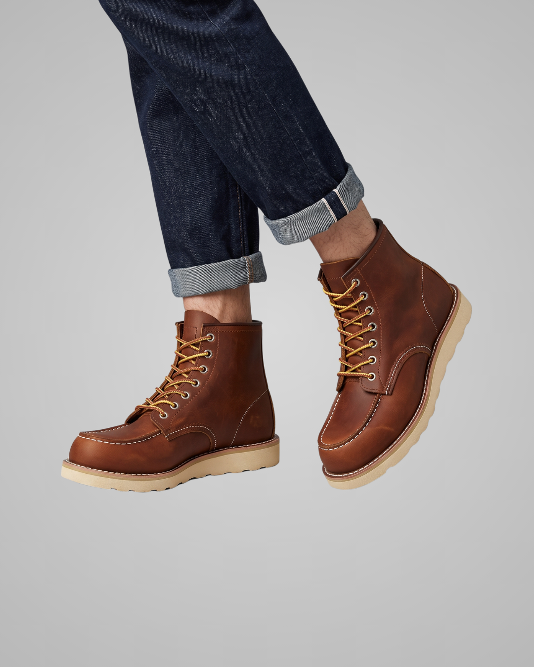 A pair of brown leather lace-up ankle boots worn with dark blue cuffed jeans, shown against a solid gray background.