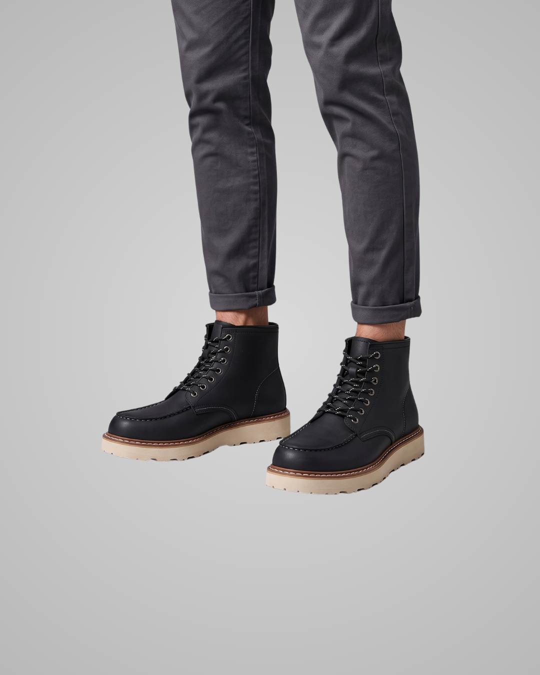 A person wearing dark gray cuffed jeans and black lace-up ankle boots with light tan soles, shown from the calves down against a plain light gray studio background.