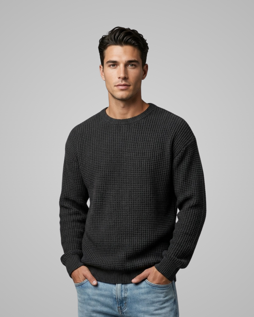 Man wearing a dark gray sweater and light blue jeans against a plain background