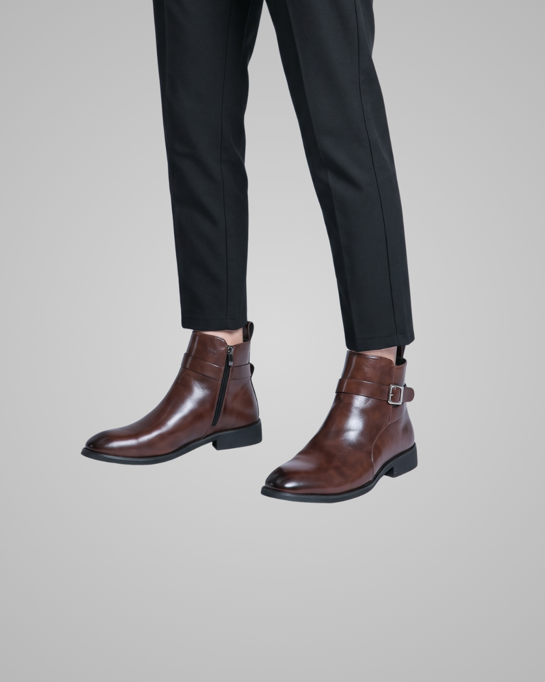 A pair of dark brown leather ankle boots with side zippers and buckle straps, shown on a model wearing black trousers against a solid gray background.