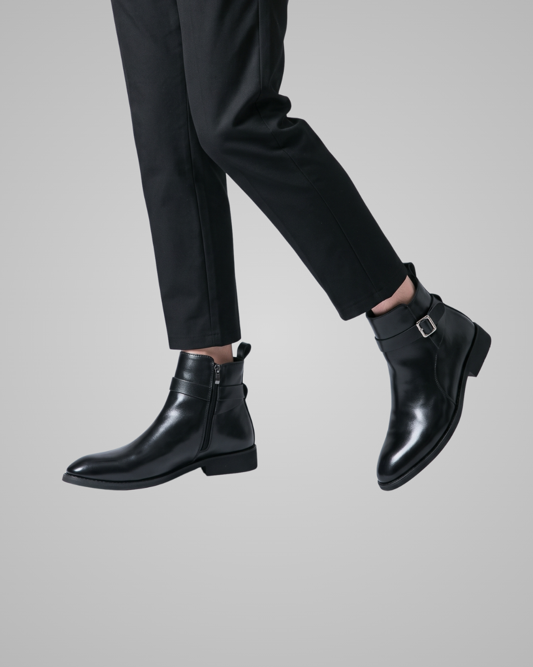 Black leather ankle boots with side zippers and buckle straps, shown on a model wearing black trousers against a solid gray background.